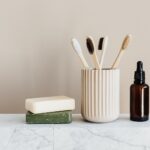 Collection of organic soaps and bamboo toothbrushes in ceramic minimalism style holder placed near renewable glass bottle with essential oil on white marble tabletop against beige wall
